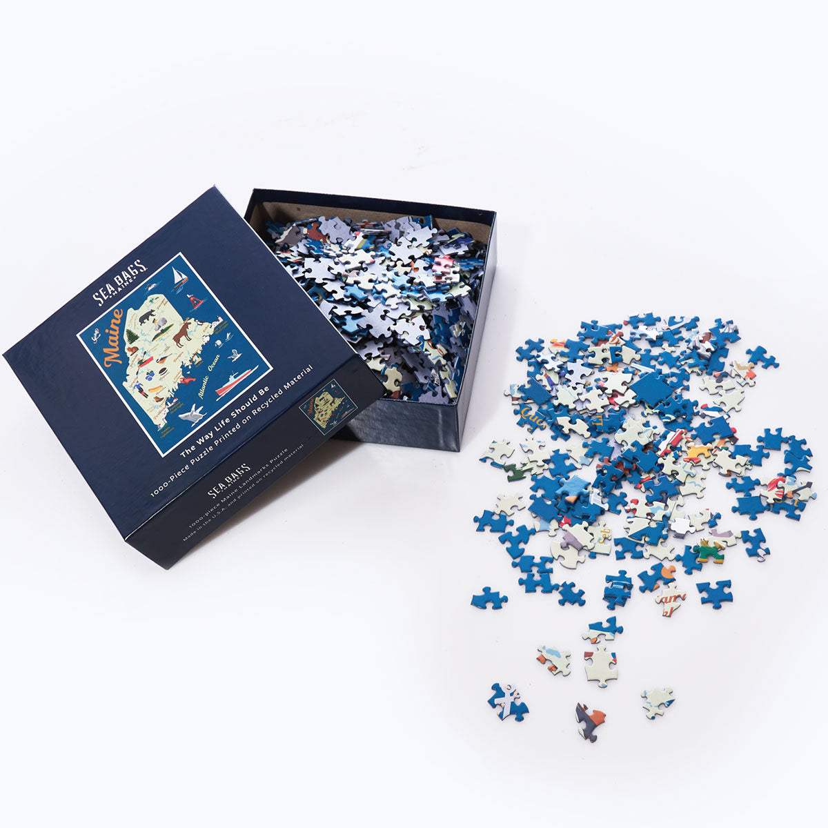A partially opened Galleyware Maine Landmarks Jigsaw Puzzle box rests on a white surface, with puzzle pieces inside and numerous blue and multicolored pieces in the shape of Maine scattered around it. by Sea Bags