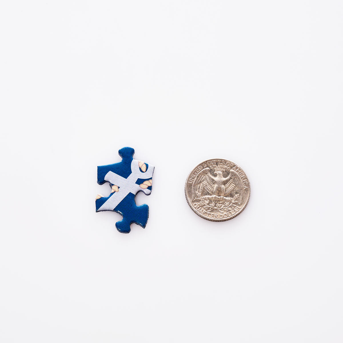 A blue and white puzzle piece from the Galleyware Maine Landmarks Jigsaw Puzzle is placed next to a U.S. quarter coin on a white background. by Sea Bags
