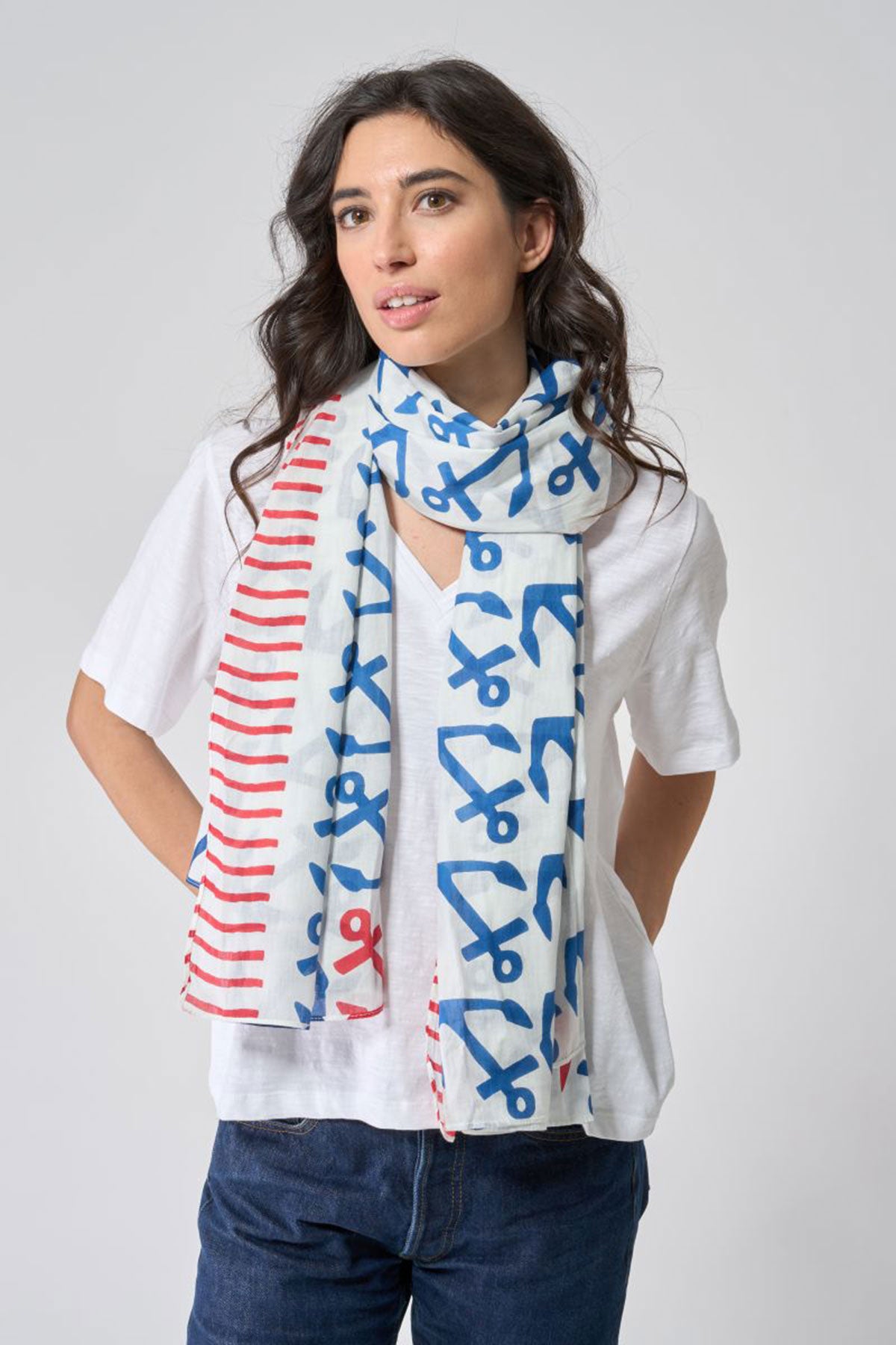 A woman stands against a plain light gray background wearing a white shirt, jeans, and the Anchor Pop Scarf by Batela USA Inc—a nautical cotton scarf adorned with blue and red scissors and stripe patterns. by Sea Bags