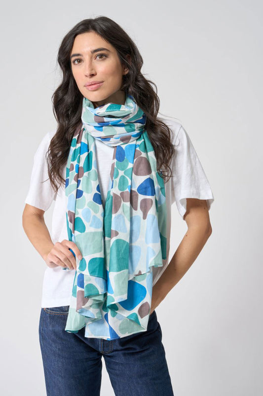 A woman with long brown hair wears a white T-shirt, blue jeans, and the Batela USA Inc Sea Glass Scarf—a light cotton accessory adorned with a blue, teal, and brown pebble-like pattern that evokes the soft hues of sea glass and brings a touch of nautical charm to her look. by Sea Bags