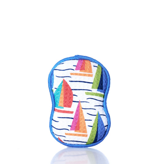 The Kate Nelligan Full Sail Dish Sponge by Once Again Home is a colorful, reusable sponge with blue trim and a cheerful sailboat pattern, crafted from recycled materials. It stands upright on a white reflective surface against a plain white background—an ideal addition to your sustainable home textiles collection. by Sea Bags