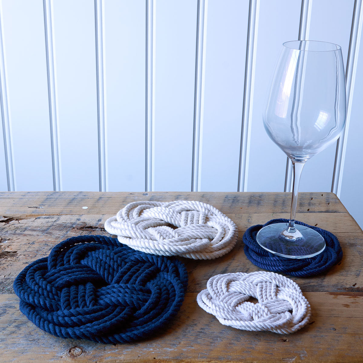 Four Nautical Sailor Knot Coasters-Natural from Mystic Knotwork, crafted in the Turk’s head knot style—two in blue and two in white—are displayed on a wooden table next to an empty wine glass.